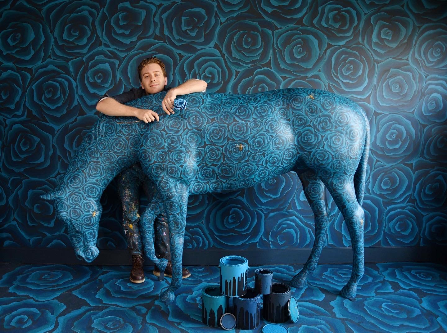 Wylie Caudill poses with a fiberglass horse painted with blue roses and gold bees in a room painted floor to ceiling in the same pattern. 